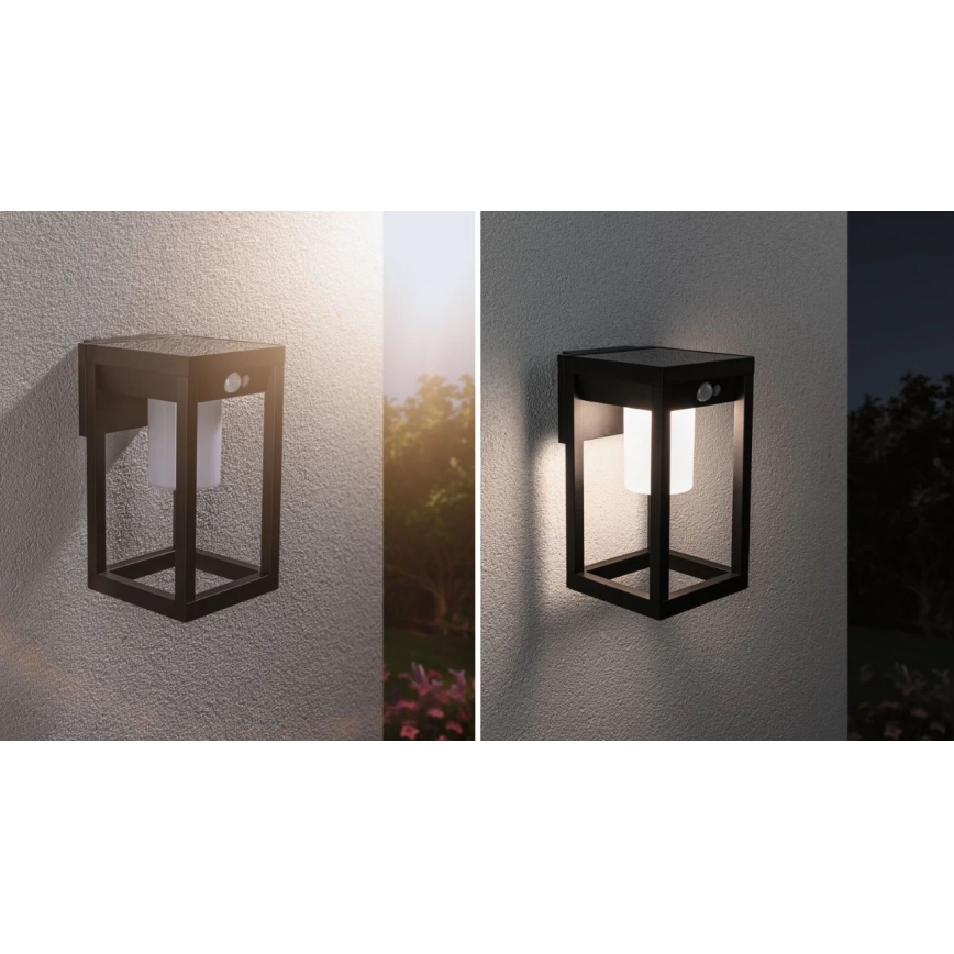 Solárna nástenná lampa s pohybovým senzorom svieti v tme. Je umiestnená na sivej stene v exteriéri. Lampa je čiernej farby, štvorcového tvaru s vnútorným mliečnym difúzorom.