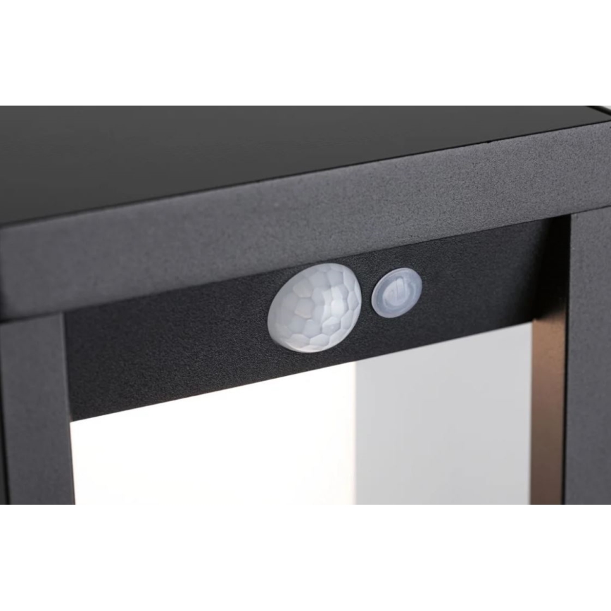 Senzor pohybu detekuje pohyb. Nachádza sa na tmavo sivej kovovej konštrukcii, pravdepodobne svietidla. Vedľa senzora je malé tlačidlo.