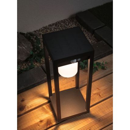 Solárna lampa svieti. Je umiestnená na drevenej terase, obklopenej kameňmi a zeleňou. Lampa má štvorcový rám a guľovité svetlo. Na vrchu je solárny panel a snímač pohybu.