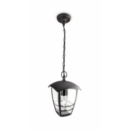 Čierna závesná lampa visí na reťazi z stropu. Lampa má štvorcový sklenený kryt a tmavý kovový kryt. Je umiestnená na bielom pozadí.