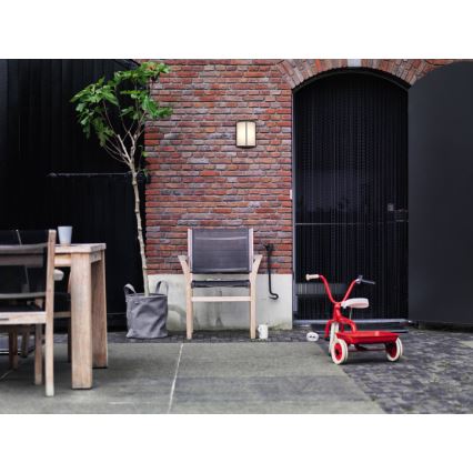 Záhradná stolička stojí na terase pred tehlovou stenou. Vedľa nej je detská trojkolka a kvetináč. Na stene je pripevnená vonkajšia lampa.