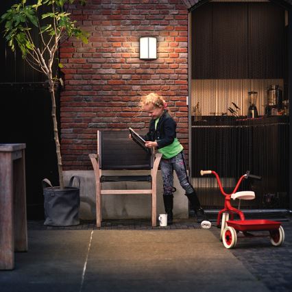 Dievča s tmavými topánkami a zelenou mikinou pri vonkajšom sedení s drevenou stoličkou prezerá knihu. Vedľa je detský trojkolka a strom v kvetináči. Kontext tvorí tehlová stena a dvere s kovovou záclonou.