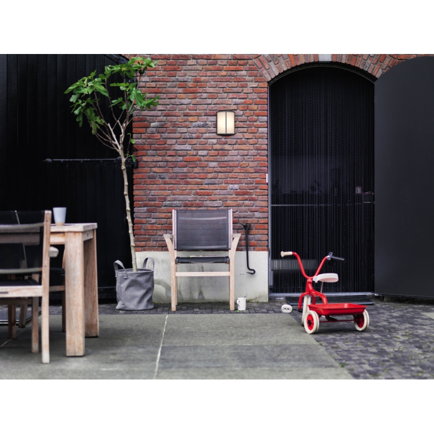 Záhradná stolička stojí na terase pred tehlovou stenou. Vedľa nej je detská trojkolka a kvetináč. Na stene je pripevnená vonkajšia lampa.