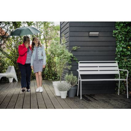 Dve ženy idú po drevenej terase pod dáždnikom. Vedľa nich je biela lavička a kvetináče s rastlinami. Na tmavej stene domu je pripevnené vonkajšie svietidlo.