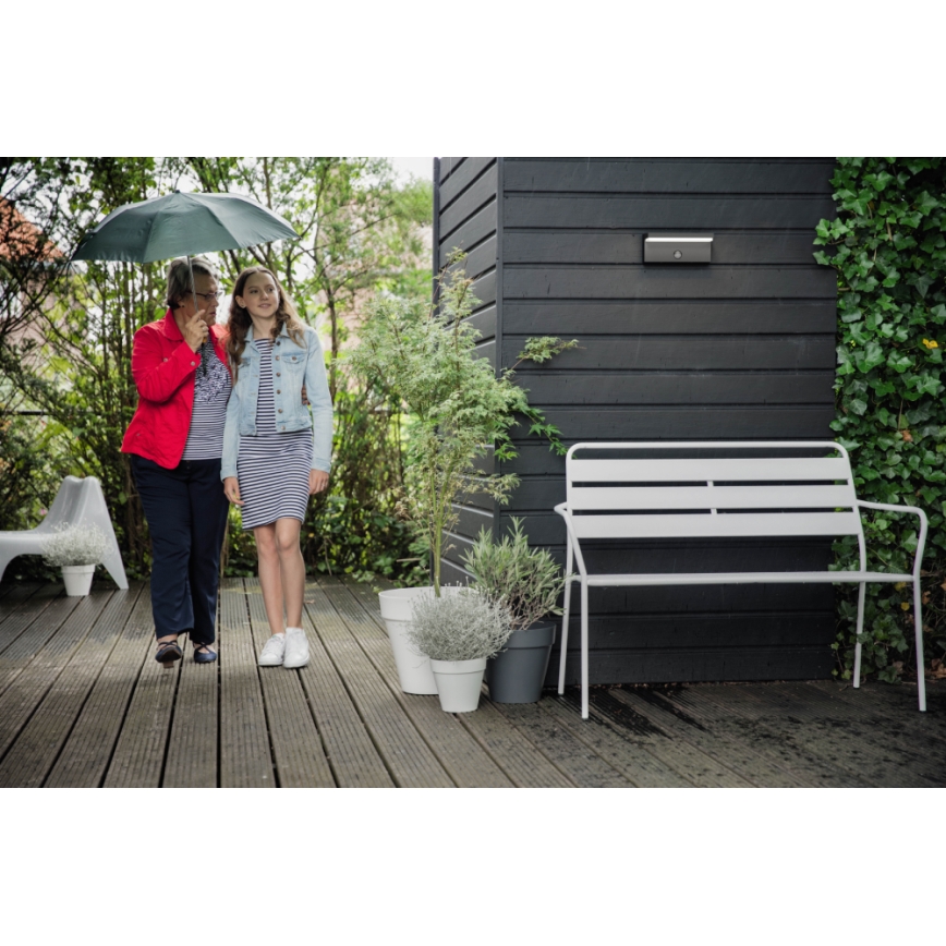 Dve ženy idú po drevenej terase pod dáždnikom. Vedľa nich je biela lavička a kvetináče s rastlinami. Na tmavej stene domu je pripevnené vonkajšie svietidlo.