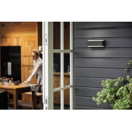 Vonkajšie nástenné svietidlo je pripevnené na tmavosivej drevenej stene. Svietidlo je obdĺžnikového tvaru. V pozadí je rozostrený interiér kuchyne s otvorenými dverami.