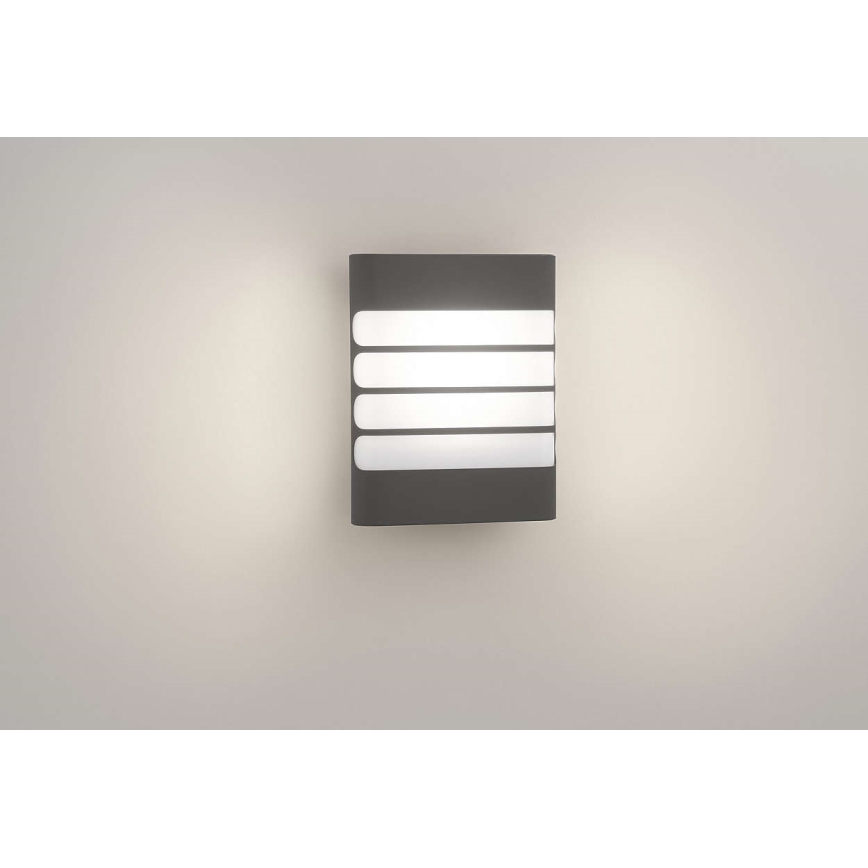 Svietidlo svieti na stene. Obdĺžnikové telo svietidla je tmavosivé, s piatimi vodorovnými bielymi prvkami, ktoré prepúšťajú svetlo. Svetlo je zapnuté.