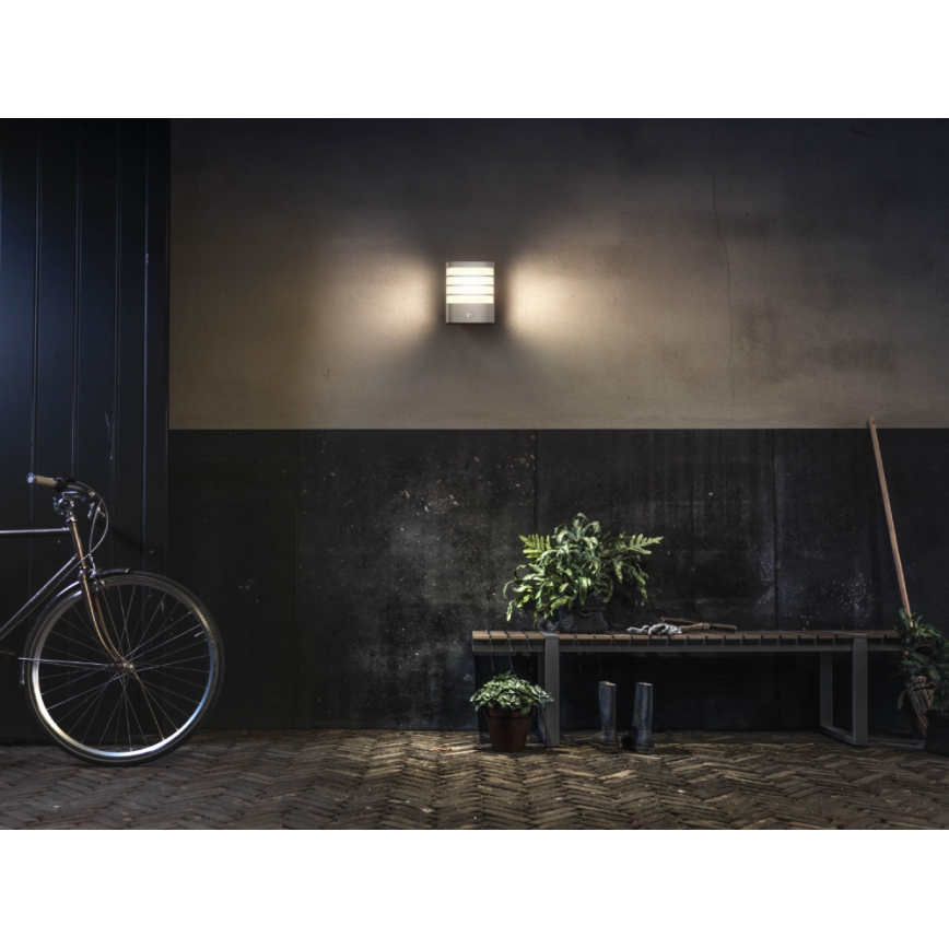 Nástenná lampa svieti v tmavšom priestore. Cyklistické kolo stojí pri stene oproti lavičke s kvetináčmi a gumákmi. Podlaha je z dlaždíc.