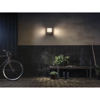 Nástenná lampa svieti v tmavej chodbe. Pri stene je bicykel, drevená lavica s kvetináčmi a gumáky. Podlaha je z dlaždíc.