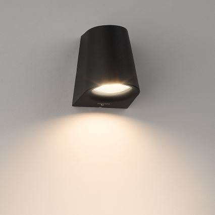Čierna nástenná lampa svieti. Lampa je pripevnená na svetlej stene. Na lampe je logo Philips.