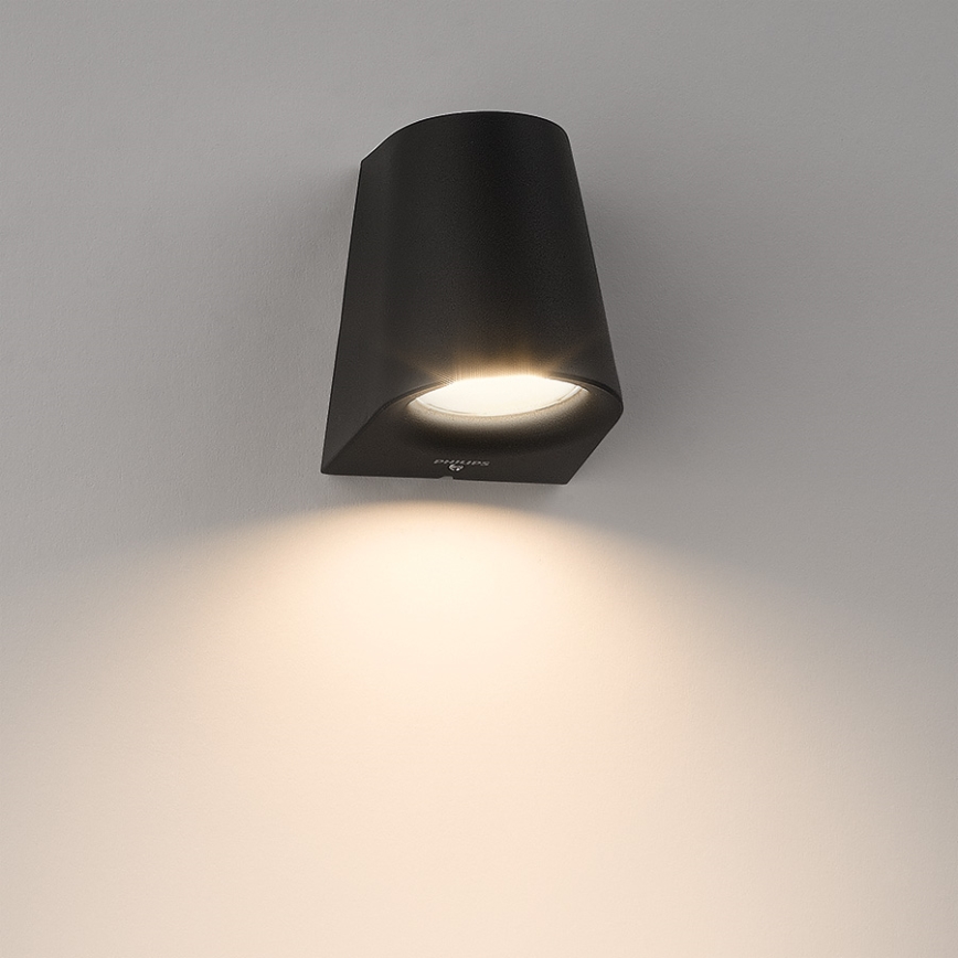 Čierna nástenná lampa svieti. Lampa je pripevnená na svetlej stene. Na lampe je logo Philips.