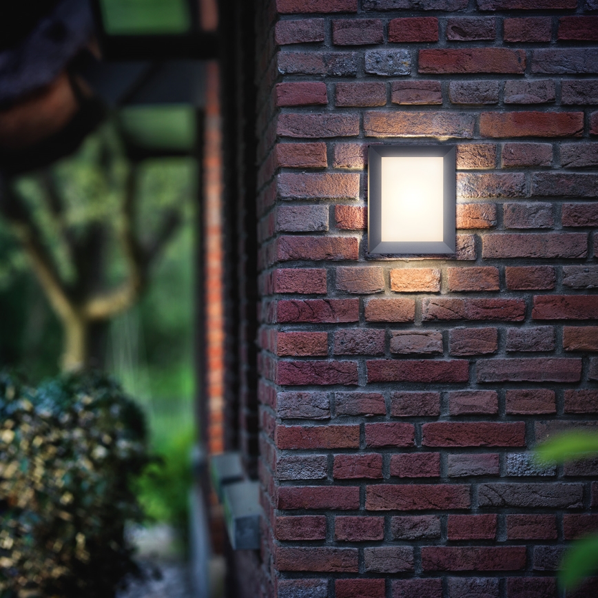 Svietidlo svieti na tehlovej stene. Svietidlo je štvorcové a zabudované do vonkajšej steny. Okolím je záhrada s kríkmi.