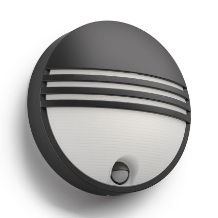 Guľatá vonkajšia nástenná lampa s integrovaným pohybovým senzorom je pripevnená na stenu. Lampa má tmavosivý rám a svetlý difúzor s priečnymi drážkami.