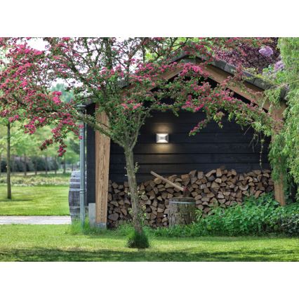 Vonkajšia nástenná lampa svieti na tmavo sfarbenú drevenú stenu hospodárskej budovy. Pri stene je naskladané drevo na kúrenie. Strom s ružovými kvetmi čiastočne zakrýva budovu.