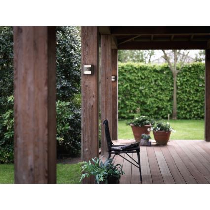 Vonkajšie nástenné svietidlo je pripevnené na drevenom stĺpe. Nachádza sa na krytej terase s drevenou podlahou, záhradným nábytkom a zeleňou.