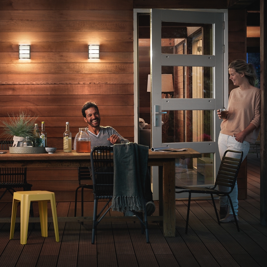 Muž a žena sedia pri stole na terase. Muž sa usmieva. Na stene sú nástenné svietidlá. Terasa je z dreva.