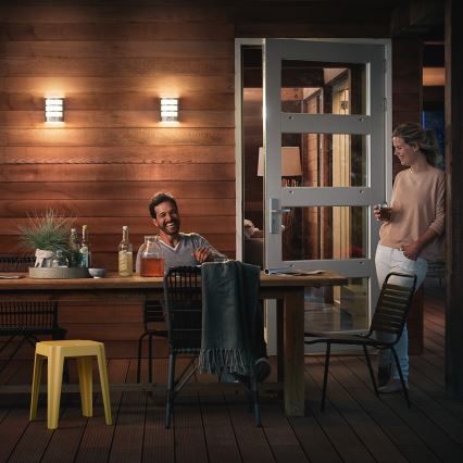 Muž a žena sedia pri vonkajšom jedálenskom stole na terase. Na stene svietia dve vonkajšie nástenné svietidlá.