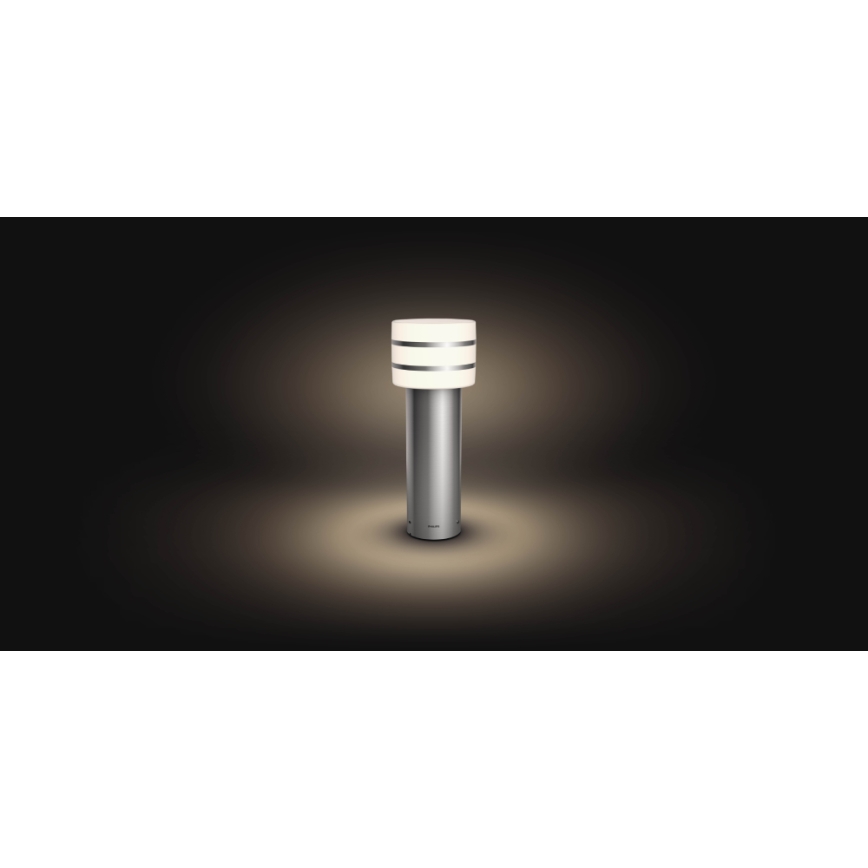 Vonkajšie svietidlo svieti na tmavom pozadí. Stĺpikové svietidlo z kovu má valcový svetelný zdroj s dvoma kovovými prstencami.