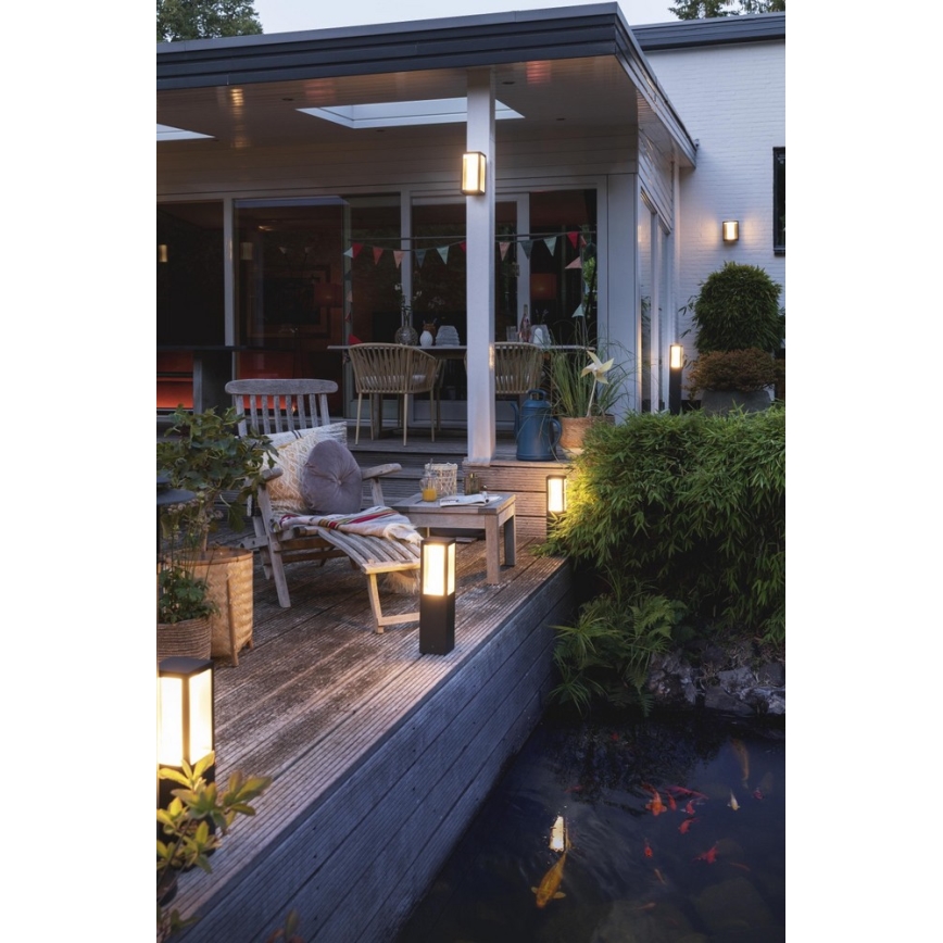 Záhradné svietidlá svietia na drevenej terase pri jazierku. Svietidlá osvetľujú posedenie s ležadlom a stolíkom. V pozadí je vidieť dom a zeleň.