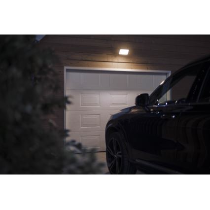 Auto stojí pred garážou osvetlenou nástenným svetlom. Garážové dvere sú biele. V popredí je hustý ker.