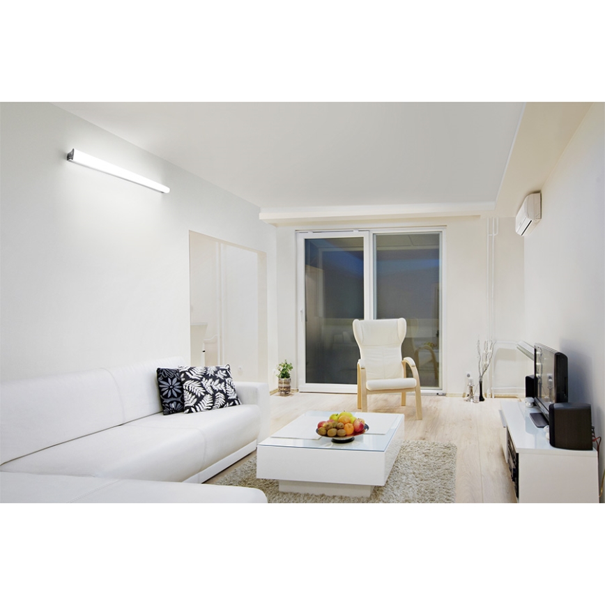 Dlhá tenká LED lampa svieti na stene obývačky. Biela sedačka a kreslo sú umiestnené v priestore s bielymi stenami, dverami a svetlou podlahou. Na konferenčnom stolíku sú ovocie.