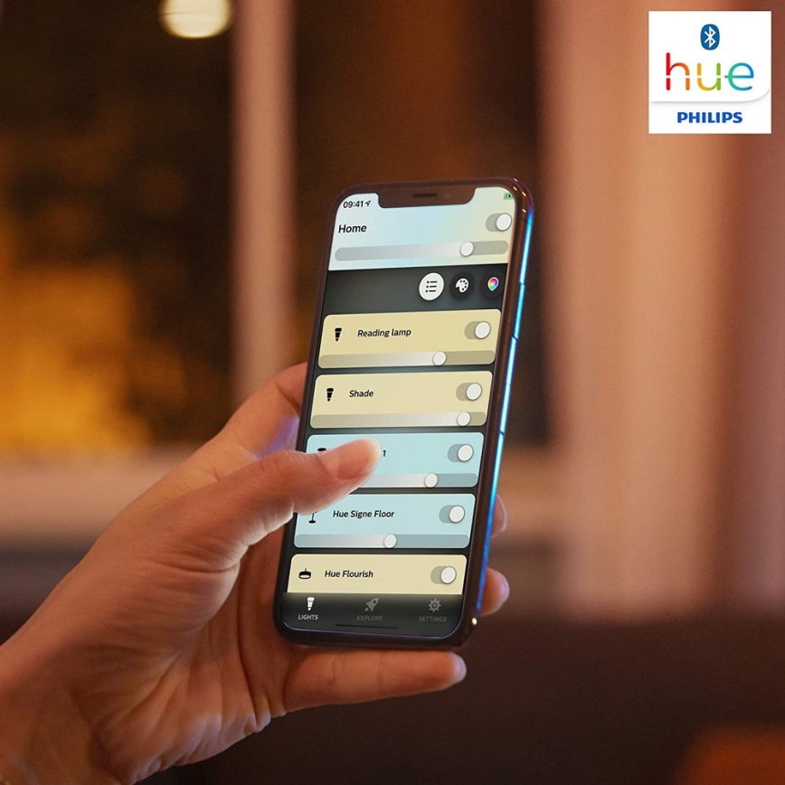 Ruka ovláda mobilný telefón s aplikáciou Philips Hue. Aplikácia zobrazuje možnosti ovládania osvetlenia: Reading lamp, Shade, Hue Signe Floor, Hue Flourish. Na pozadí je rozmazané prostredie interiéru.