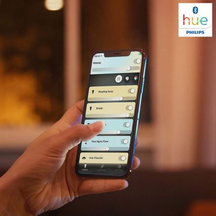 Ruka ovláda mobilný telefón s aplikáciou Philips Hue. Aplikácia zobrazuje ovládanie osvetlenia: Reading lamp, Shade, Hue Signe Floor, Hue Flourish. Prostredie je zatienená izba.