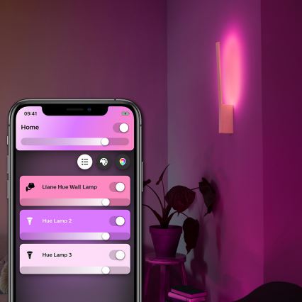 Mobilný telefón ovláda inteligentné osvetlenie. Na displeji sa zobrazuje aplikácia s názvami „Liane Hue Wall Lamp“, „Hue Lamp 2“ a „Hue Lamp 3“ a posuvníkmi na nastavenie intenzity svetla. Na stene svieti ružovo-fialová nástenná lampa.