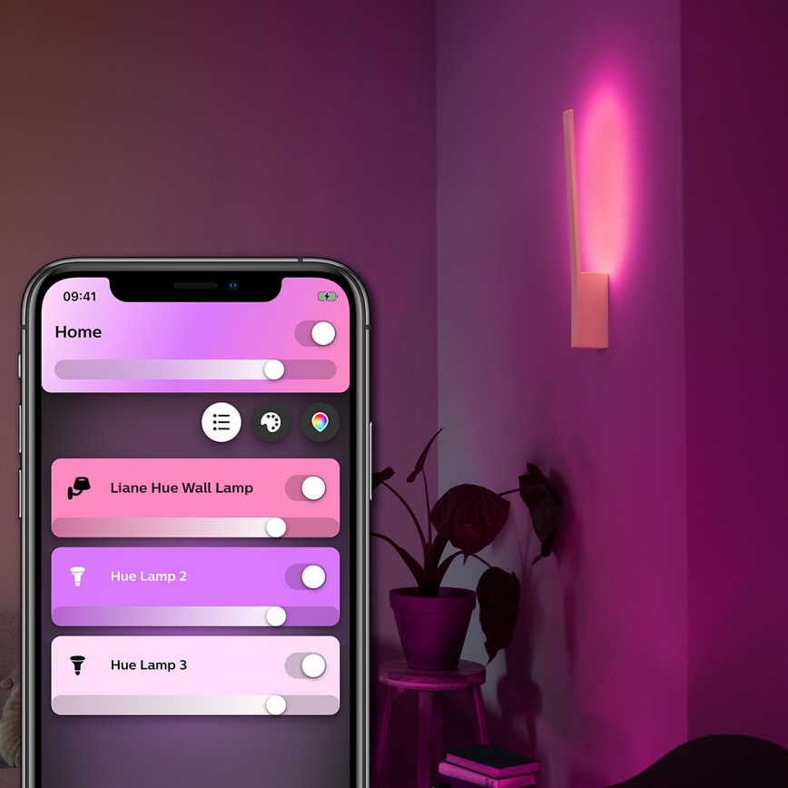 Mobilný telefón ovláda inteligentné osvetlenie. Na displeji sa zobrazuje aplikácia s názvami „Liane Hue Wall Lamp“, „Hue Lamp 2“ a „Hue Lamp 3“ a posuvníkmi na nastavenie intenzity svetla. Na stene svieti ružovo-fialová nástenná lampa.