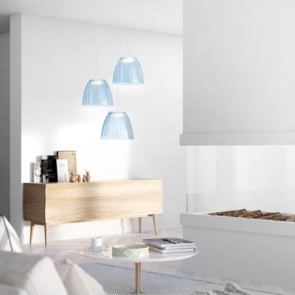 Tri svetlomodré sklenené závesné lampy s rebrovaným povrchom visia v svetlej obývacej izbe nad drevenou komodou. V pozadí sa nachádza moderný biely krb.