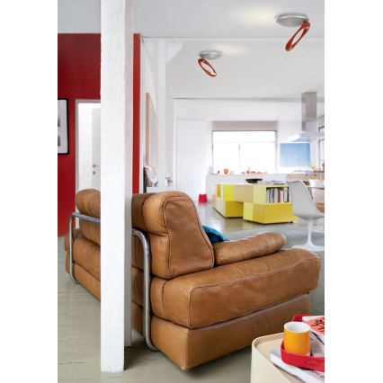 Kožená sedačka s kovovou konštrukciou stojí v otvorenom priestore. Sedačka je hnedá. V pozadí je vidieť kuchyňu a žlté skrine. Na strope sú dve stropné svietidlá s oranžovými detailmi.