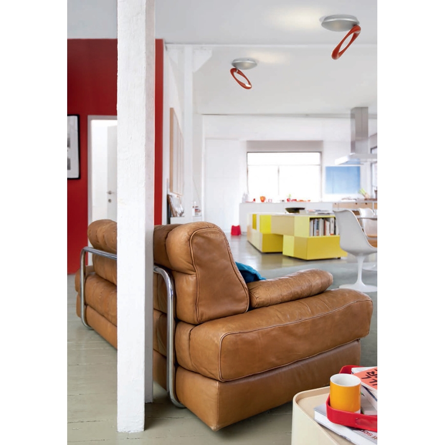 Kožená sedačka s kovovou konštrukciou stojí v otvorenom priestore. Sedačka je hnedá. V pozadí je vidieť kuchyňu a žlté skrine. Na strope sú dve stropné svietidlá s oranžovými detailmi.