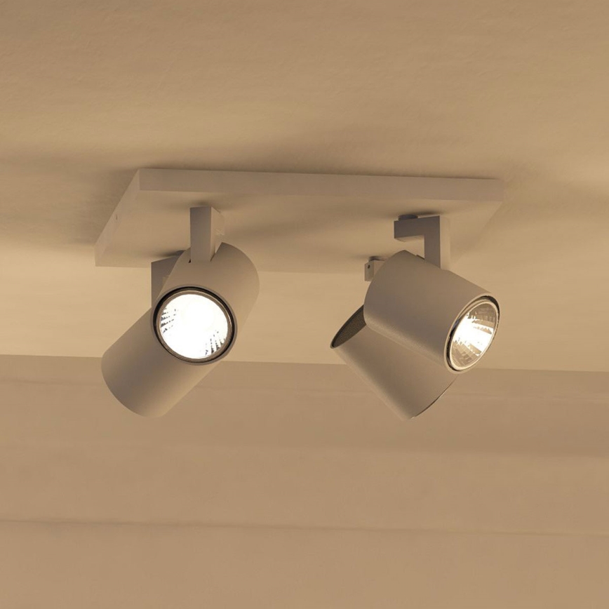 Dvojité stropné svietidlo s valcovými tienidlami svieti na svetlo. Svietidlá sú pripevnené k obdĺžnikovej základni pripevnenej na strope. Svetlo je teplej farby.