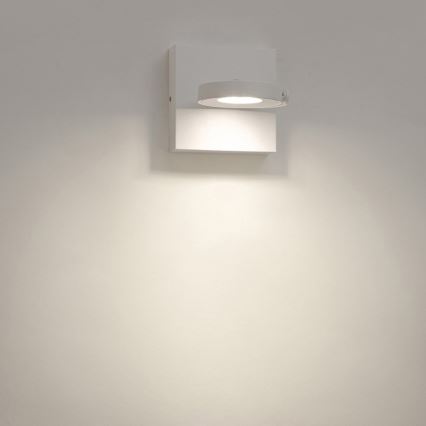 Biela nástenná lampa svieti na stenu. Lampa je pripevnená k bielej stene.