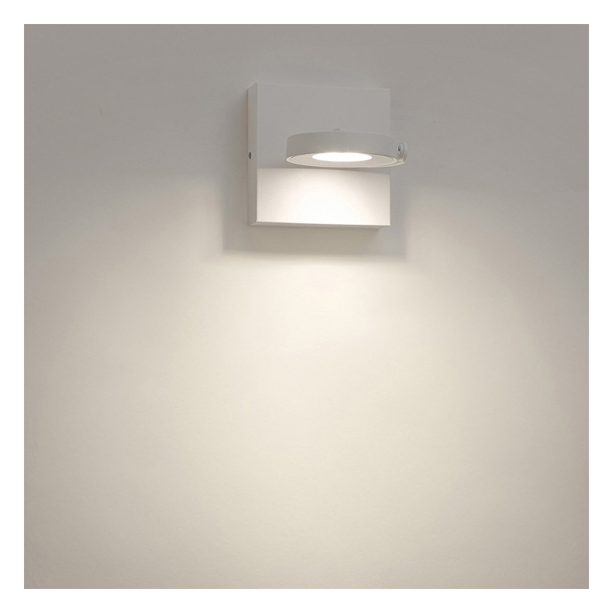Biela nástenná lampa svieti na stenu. Lampa je pripevnená k bielej stene.