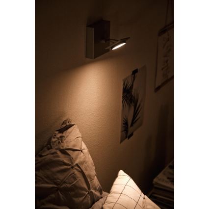 Nástenná lampa svieti na posteľ. Lampa je pripevnená na stenu v spálni. V pozadí sú viditeľné obrazy a vankúše na posteli.