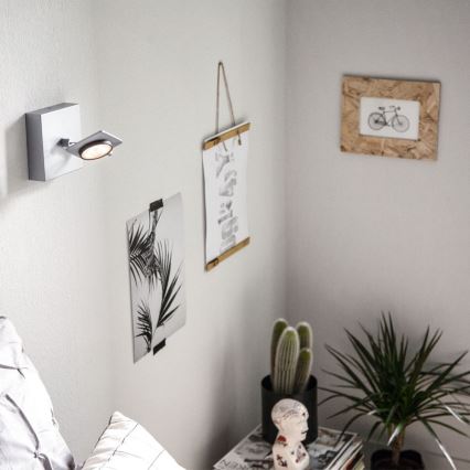 Svietidlo je pripevnené na stene. Svieti smerom nadol. Na stene sú zavesené obrázky a rastliny sú na nočnom stolíku.