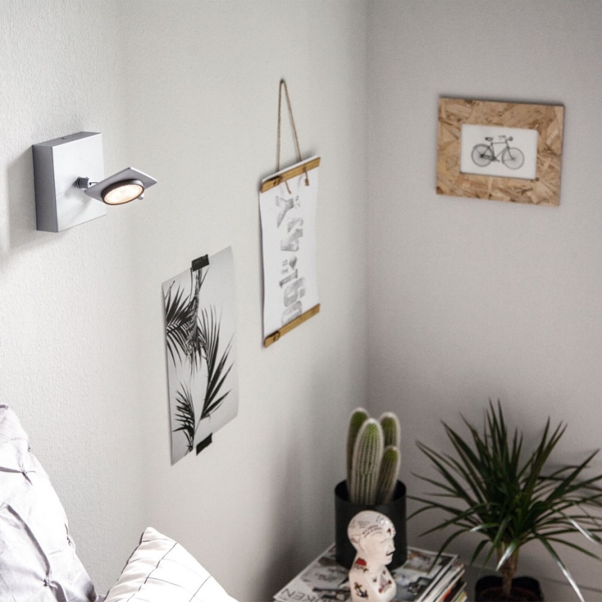 Svietidlo je pripevnené na stene. Svieti smerom nadol. Na stene sú zavesené obrázky a rastliny sú na nočnom stolíku.