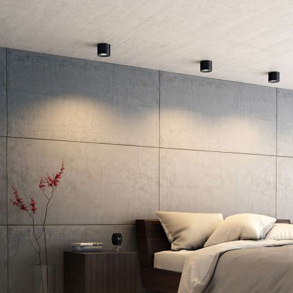 Svietidlá svietia v spálni. Tri vstavané stropné svietidlá osvetľujú stenu a posteľ s posteľnou bielizňou. Stena je z betónu.