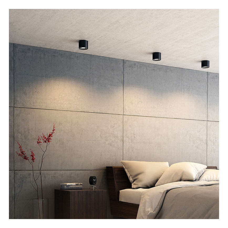 Svietidlá svietia v spálni. Tri vstavané stropné svietidlá osvetľujú stenu a posteľ s posteľnou bielizňou. Stena je z betónu.