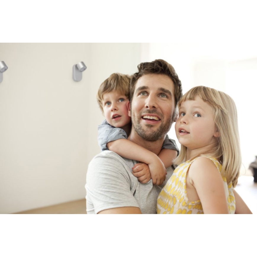 Otec drží dve malé deti na pleciach. Deti sa pozerajú nahor. V pozadí sú viditeľné dve nástenné svietidlá.