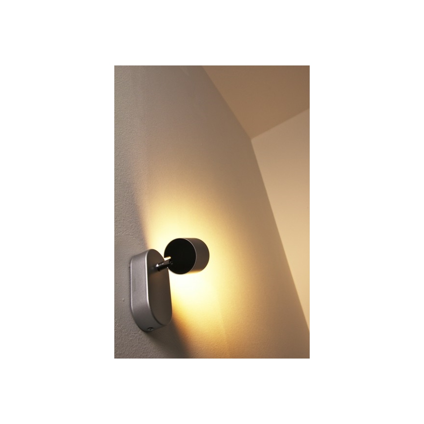 Strieborná nástenná lampa svieti na svetlošedkú stenu. Lampa je otočná, jej hlavica smeruje doprava. Svetlo je teplé, žlté.