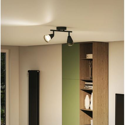 Dvojramenné stropné svietidlo svieti v interiéri. Svietidlo je čierne, s dvoma nastaviteľnými reflektormi. Nachádza sa v miestnosti s knižnicou a radiátorom.