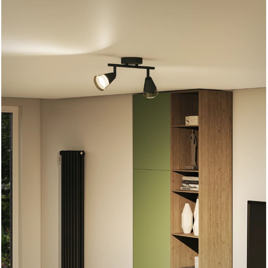 Dvojramenné stropné svietidlo svieti v interiéri. Svietidlo je čierne, s dvoma nastaviteľnými reflektormi. Nachádza sa v miestnosti s knižnicou a radiátorom.