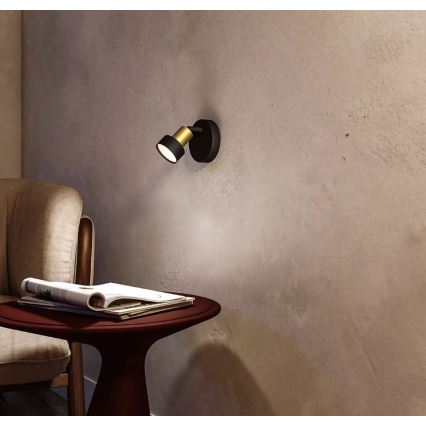 Čierna nástenná lampa so zlatou časťou svieti. Lampa je umiestnená na svetlohnedej stene popri hnedom kresle a červenom stolíku s časopismi.