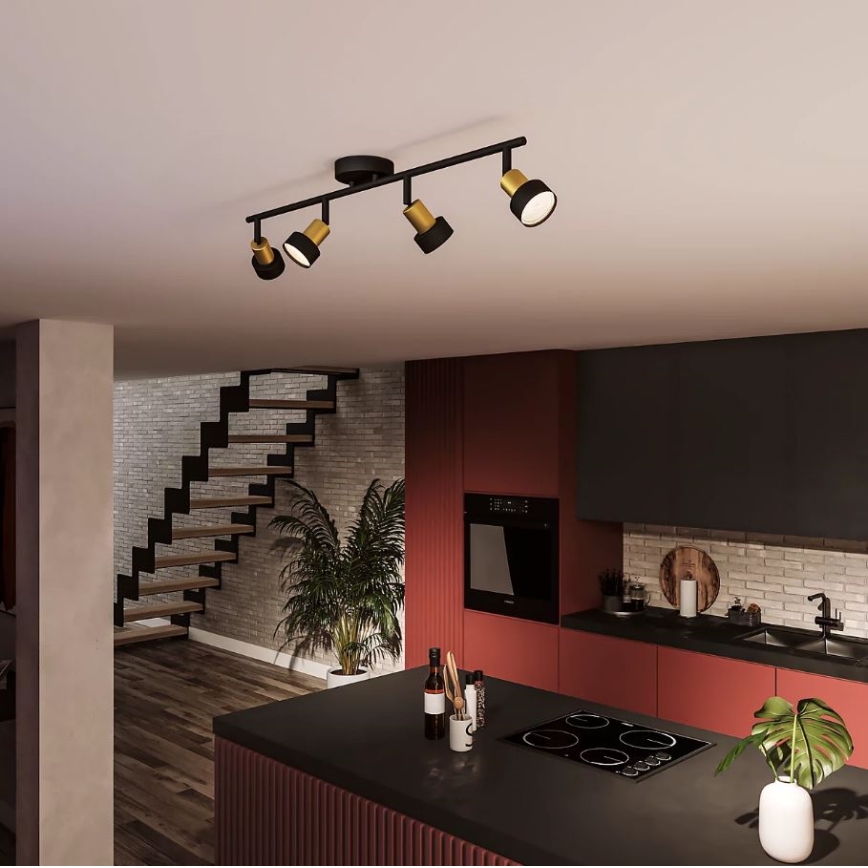 Čtveřice reflektorů s černými stínidly a zlatými detaily je namontována na stropní liště v moderní kuchyni. Lišta je černá. Kuchyň obsahuje ostrov s varnou deskou, dřevenou podlahu a schody v pozadí.