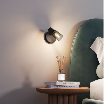 Nástenná lampa svieti v spálni. Je pripevnená na stene vedľa nočného stolíka, na ktorom sú knihy s názvami „HOTEL DESIGN“ a „The Modern Sunset“ a difuzér.