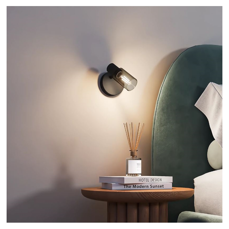 Nástenná lampa svieti v spálni. Je pripevnená na stene vedľa nočného stolíka, na ktorom sú knihy s názvami „HOTEL DESIGN“ a „The Modern Sunset“ a difuzér.