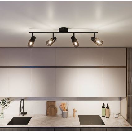 Čtyřramenné stropní svítidlo s černými kovovými rameny a válcovými skleněnými difúzory svítí v kuchyni nad kuchyňskou linkou s dřezem a varnou deskou.