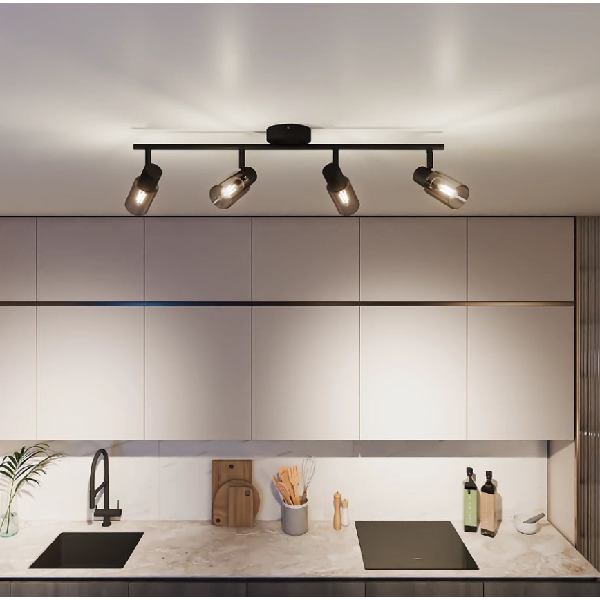 Čtyřramenné stropní svítidlo s černými kovovými rameny a válcovými skleněnými difúzory svítí v kuchyni nad kuchyňskou linkou s dřezem a varnou deskou.